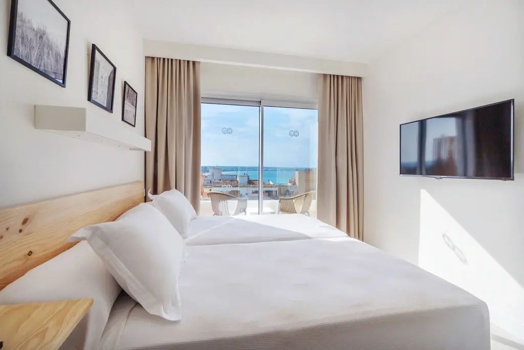Doppelbett mit weißer Bettwäsche und Blick durch eine Glasfensterfront auf das Meer.