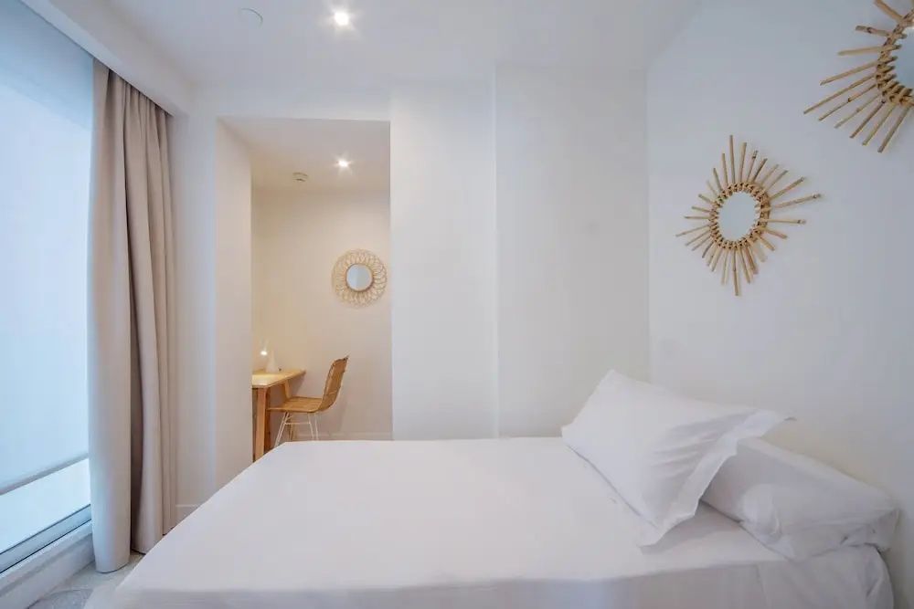 Weiß eingerichtetes Doppelzimmer mit sonnenförmigem Wandspiegel und kleinem Schreibtisch im Hintergrund.