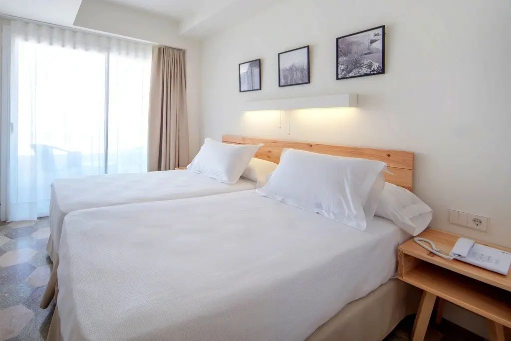 Doppelbett mit weißer Bettwäsche und Kopfteil aus Holz in einem hellen Hotelzimmer.