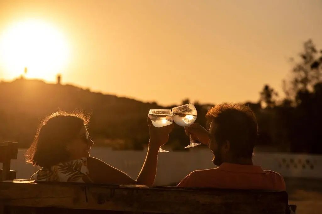 Zwei Personen stoßen bei Sonnenuntergang auf einer Terrasse mit Gläsern an.