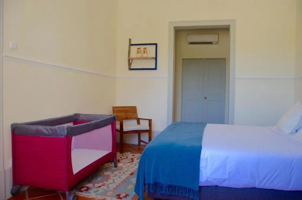 Schlafzimmer mit Doppelbett, rosa Reisebettchen für Kinder und Stuhl auf Fliesenboden.