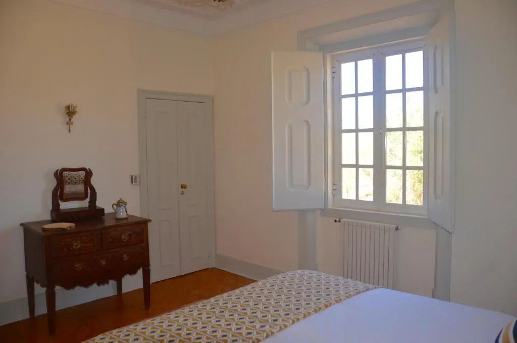 Hell erleuchtetes Zimmer mit Bett, Holzkommode und einem Fenster mit weißem Klappladen.