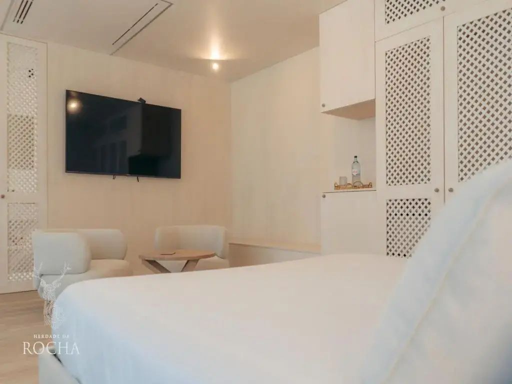 Helles Hotelzimmer mit großem Doppelbett, Fernseher an der Wand und kleinen Sesseln.