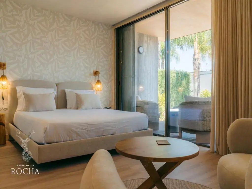 Helles Hotelzimmer mit Doppelbett, kleinem Holztisch und Glasfront zur überdachten Terrasse.