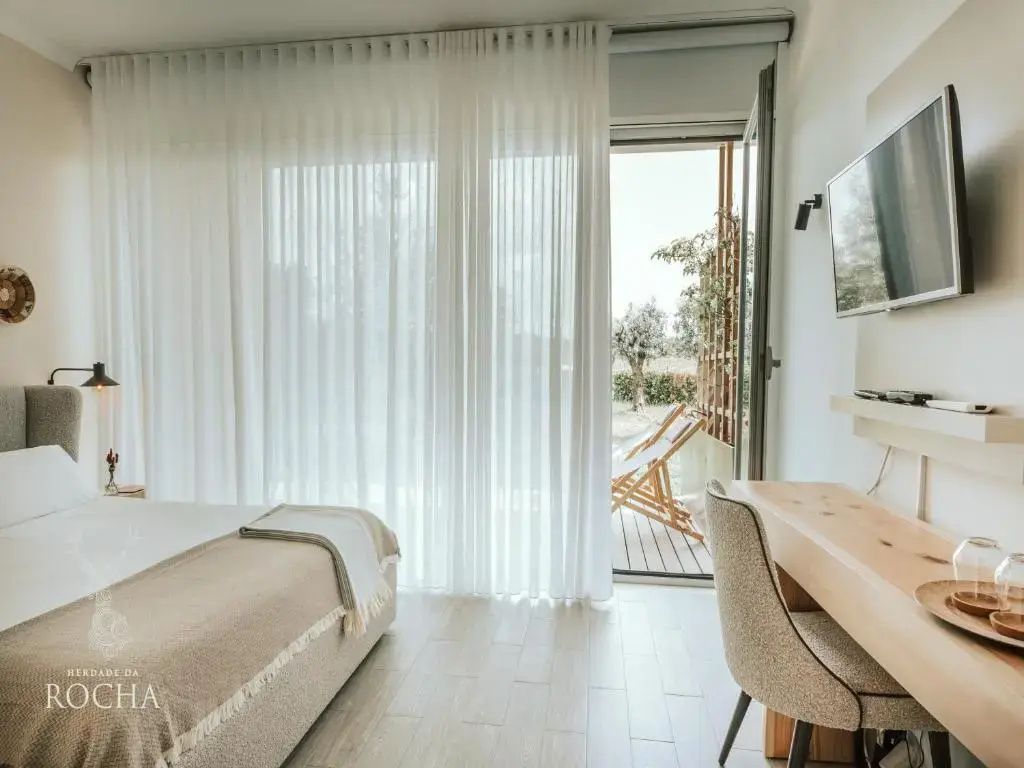 Helles Hotelzimmer mit Schreibtisch und offenem Zugang zu einer Holzterrasse mit Liegestühlen.