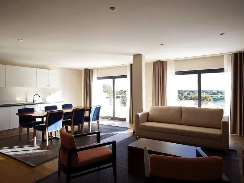Geräumiger Wohnbereich mit beigem Sofa, Sesseln und einem Esstisch mit blauen Stühlen.
