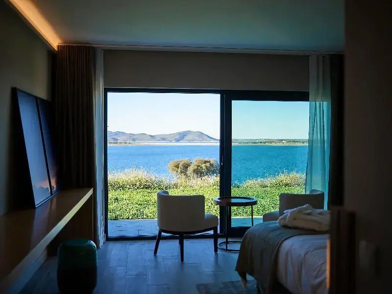 Blick aus dem Hotelzimmer durch eine große Glasschiebetür auf den See und Hügel.
