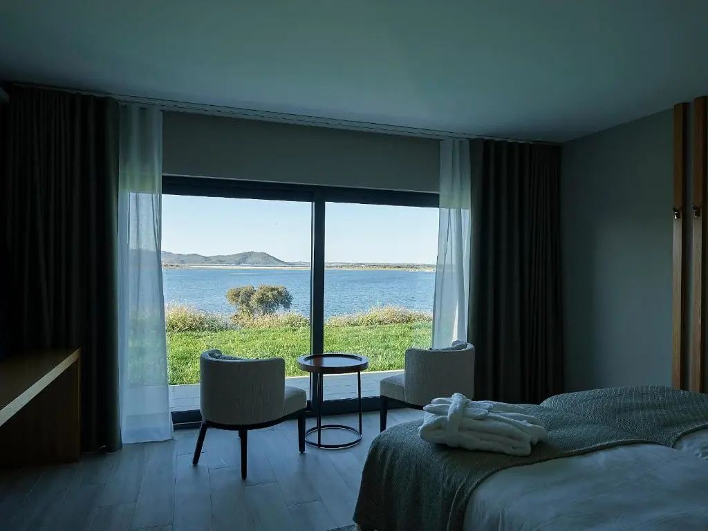 Blick durch das große Panoramafenster im Zimmer auf den See und grüne Wiese.