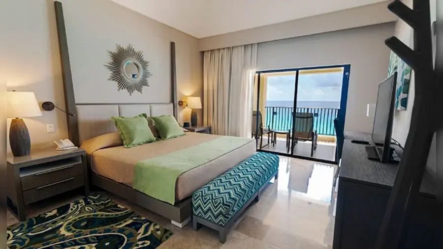 Hotelzimmer mit großem Doppelbett, grünen Kissen, Sitzbank und Balkon mit Meerblick.