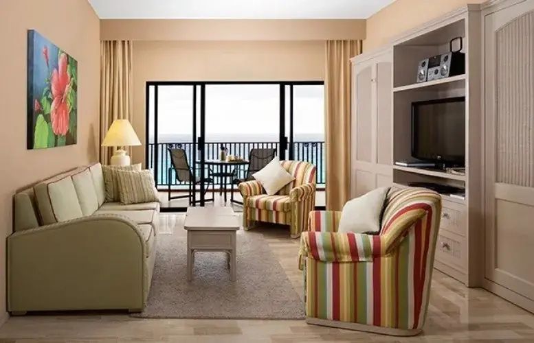 Wohnbereich mit gestreiften Sesseln, Sofa, Fernseher und Balkon mit Blick auf das Meer.