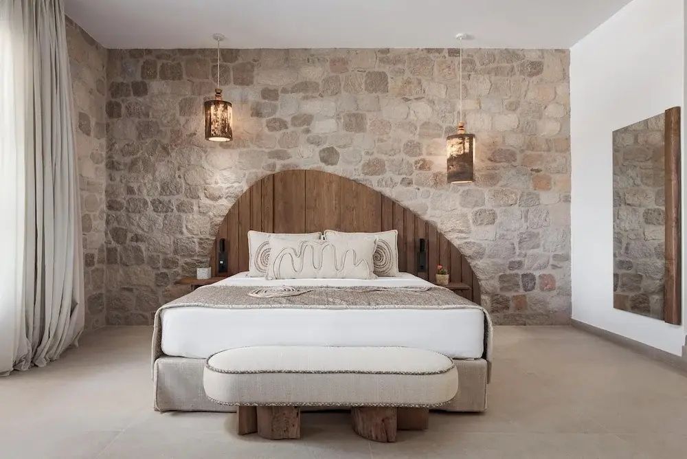 Doppelbett mit Holzkopfteil vor einer Natursteinwand und zwei hängenden Lampen.