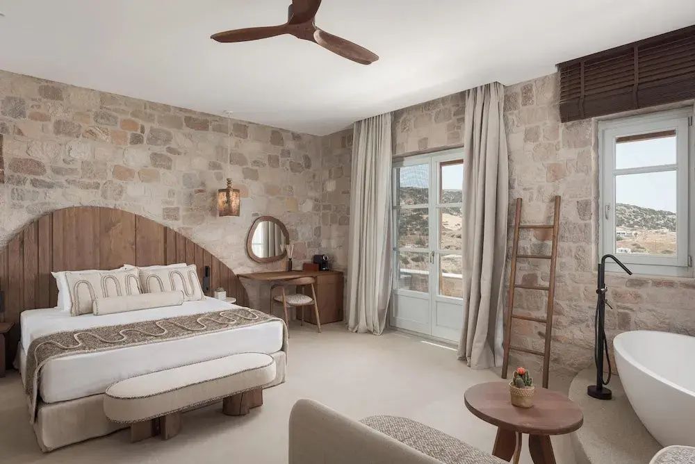 Schlafbereich mit Doppelbett, Natursteinwand, Holzventilator und Balkontür ins Freie.