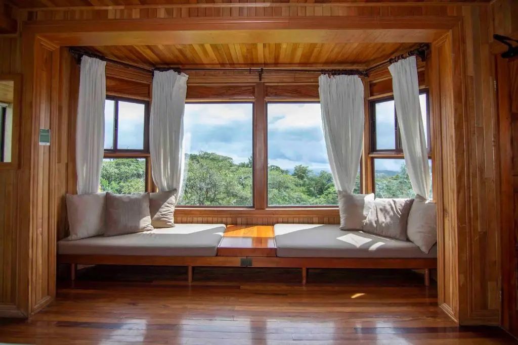 Fensterfront aus Holz mit gepolsterter Sitzbank und Ausblick auf den grünen Wald.