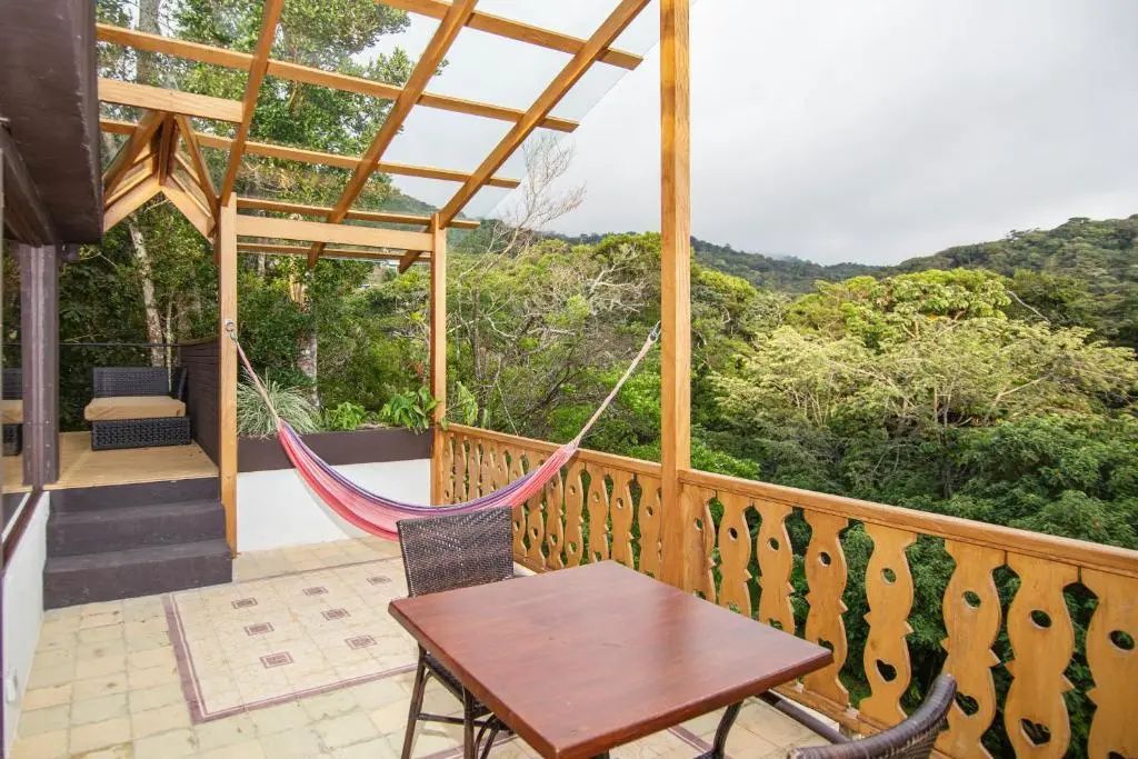 Balkon aus Holz mit Hängematte, Sitzbereich und Aussicht auf dicht bewaldete Hügel.