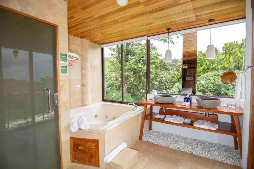 Badezimmer mit einer Whirlpoolwanne und zwei Steinwaschbecken vor Fenstern im Grünen.