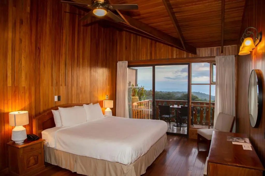 Holzverkleidetes Hotelzimmer mit Doppelbett, Deckenventilator und Glasflügeltür zum Balkon.