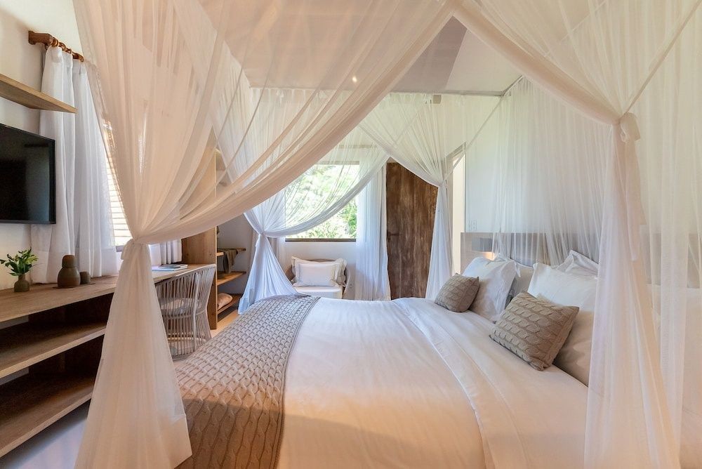 Hell eingerichtetes Hotelzimmer mit einem Himmelbett, Holzmöbeln und einem Sessel am Fenster.