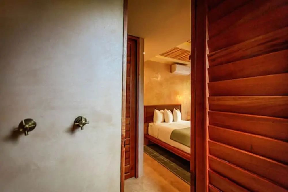 Blick durch eine geöffnete Holztür in ein Hotelzimmer mit Doppelbett und Deckenventilator.