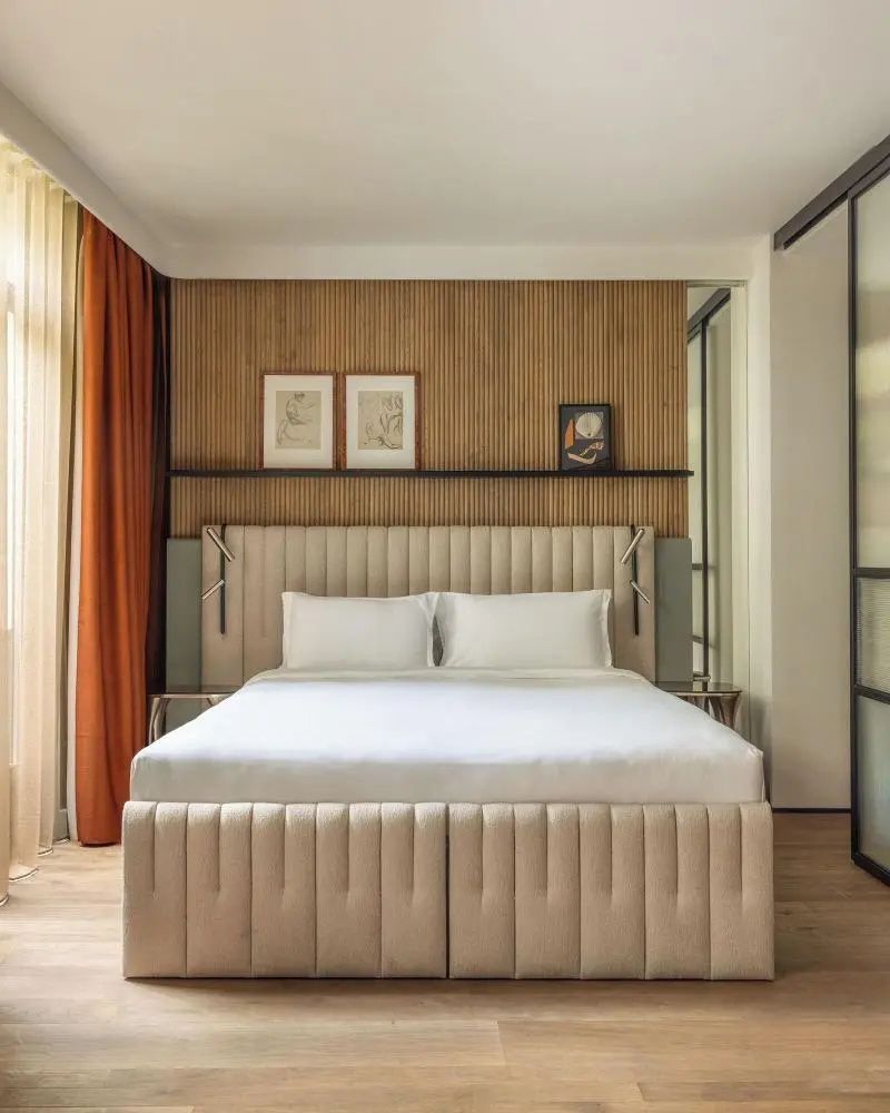 Doppelbett mit hellbeigem Polsterkopfteil vor Holzvertäfelung und Wandbildern im Hotelzimmer.