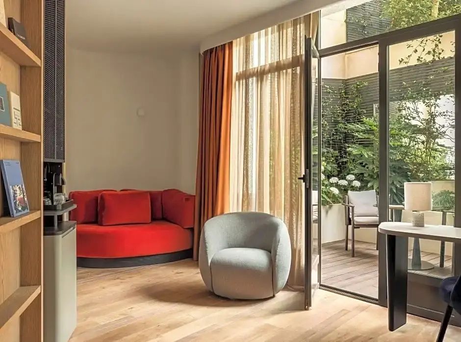 Sitzbereich im Hotelzimmer mit rotem Rundsofa, grauem Sessel und Zugang zur begrünten Terrasse.