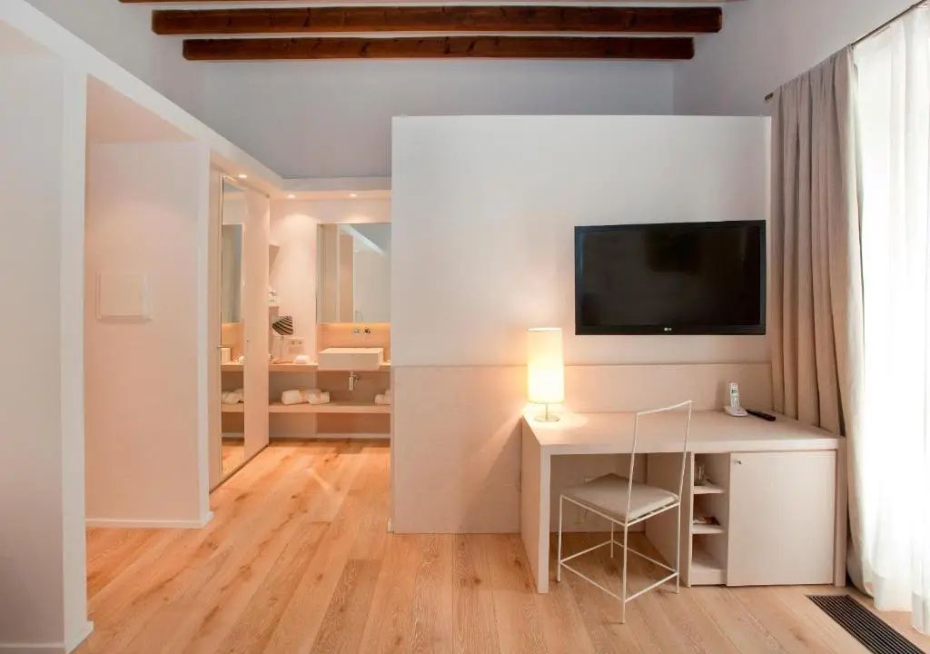 Helles Hotelzimmer mit Holzboden, Schreibtisch, Flachbildfernseher und Blick ins Badezimmer.
