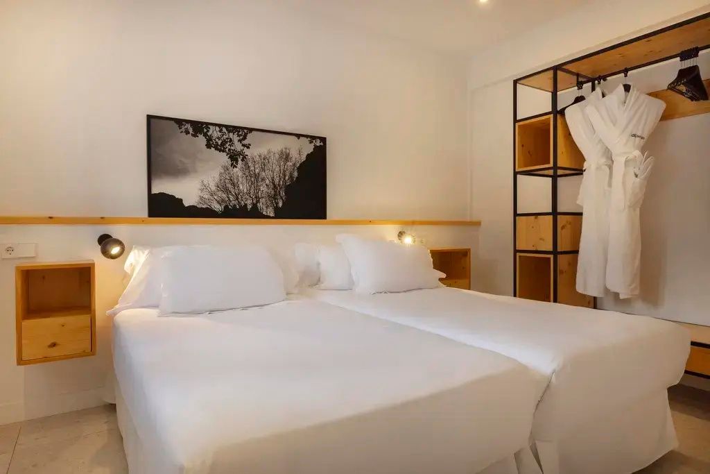 Doppelbett mit weißer Bettwäsche und ein offener Kleiderschrank im hellen Hotelzimmer.