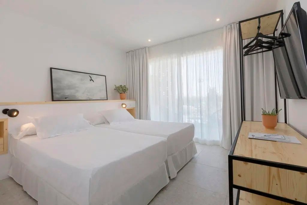 Hell eingerichtetes Hotelzimmer mit zwei Betten, weißer Bettwäsche und Vorhängen vor dem Fenster.