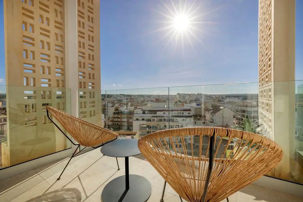 Sonniger Balkon mit zwei Flechtstühlen, einem Beistelltisch und Blick über die Stadt.