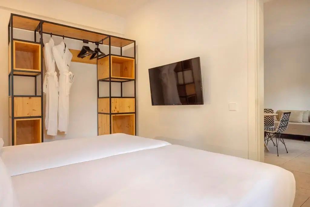 Ein Wandfernseher gegenüber einem weißen Bett und ein offenes Regalsystem für Kleidung.