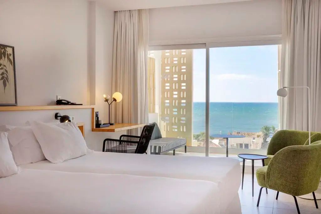 Heller Hotelraum mit Doppelbett, Schreibtisch und großem Fenster mit Blick auf das Meer.