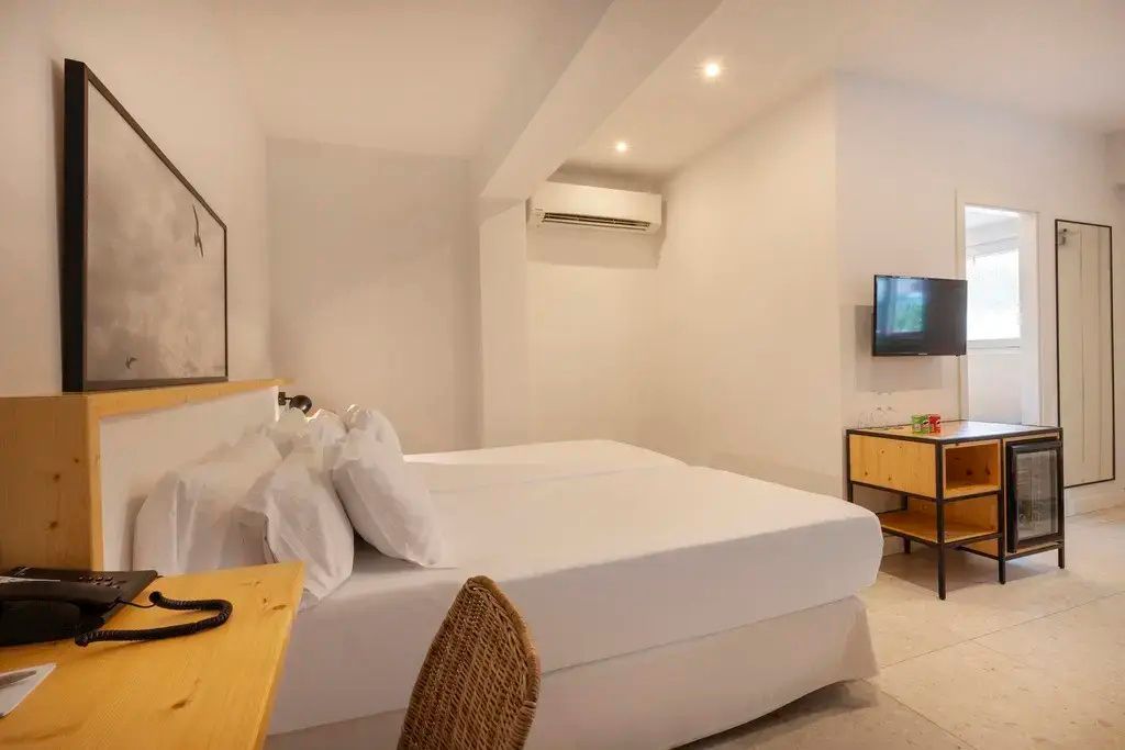 Doppelbett mit weisser Bettwaesche, Klimaanlage an der Wand und Fernseher.