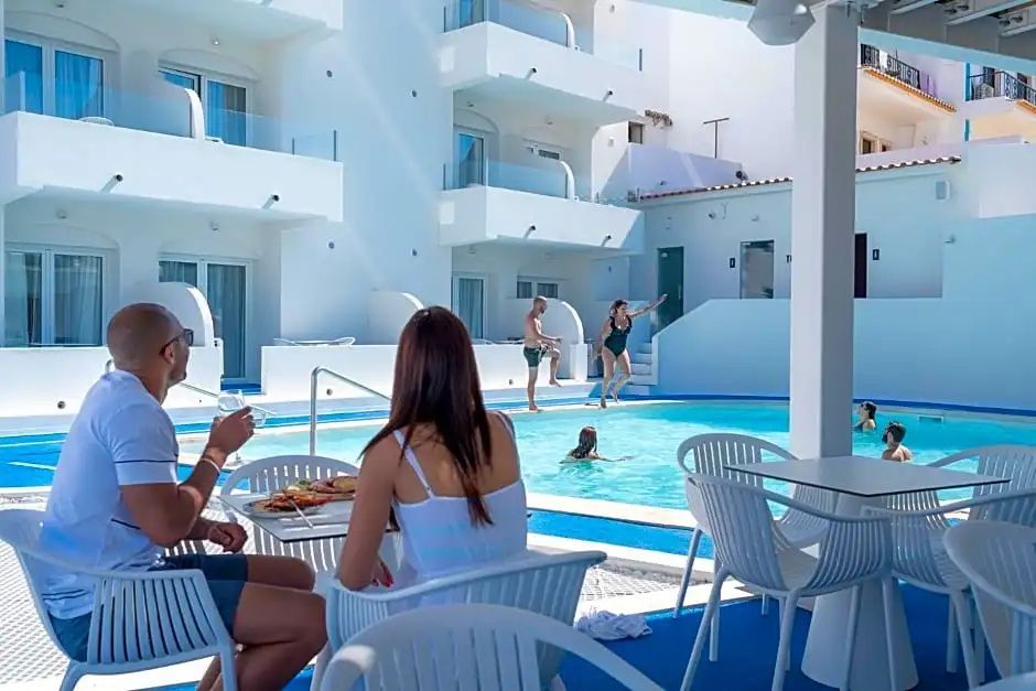 Gäste sitzen an Tischen auf einer Terrasse neben einem Pool vor den weißen Hotelgebäuden.