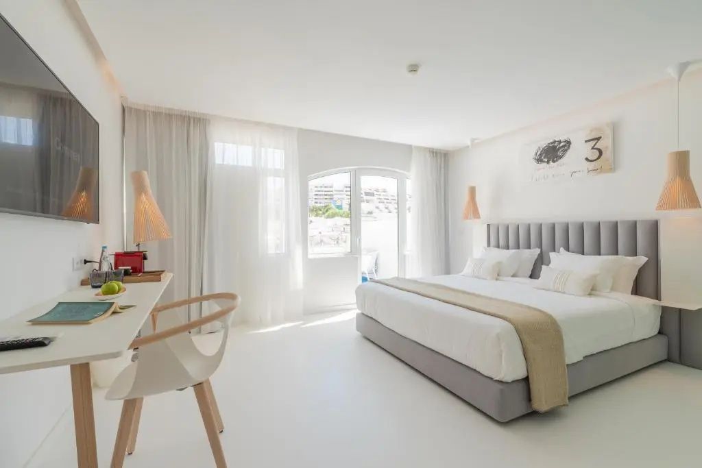Lichtdurchflutetes Zimmer mit großem Doppelbett, weißem Schreibtisch und Balkontür.