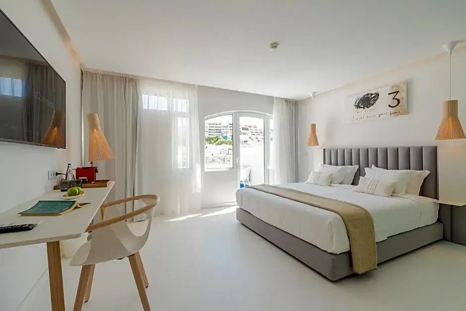 Helles Doppelzimmer mit grauem Bett, Schreibtisch und großem Fenster mit weißen Gardinen.