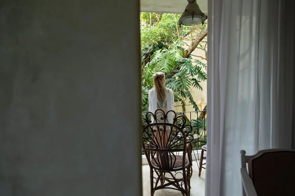 Eine Frau steht auf einem Balkon mit Rattanstühlen, umgeben von dichten grünen Pflanzen.