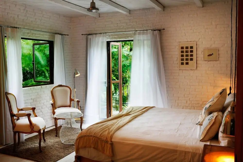 Schlafzimmer mit weiß getünchten Backsteinwänden, Doppelbett und geöffneter Balkontür ins Grüne.