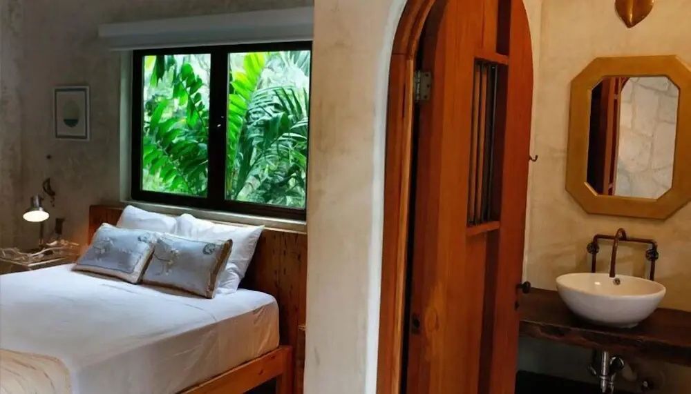Ein Hotelzimmer mit Holzbett, weißer Bettwäsche und Fenster mit Blick auf tropische Pflanzen.