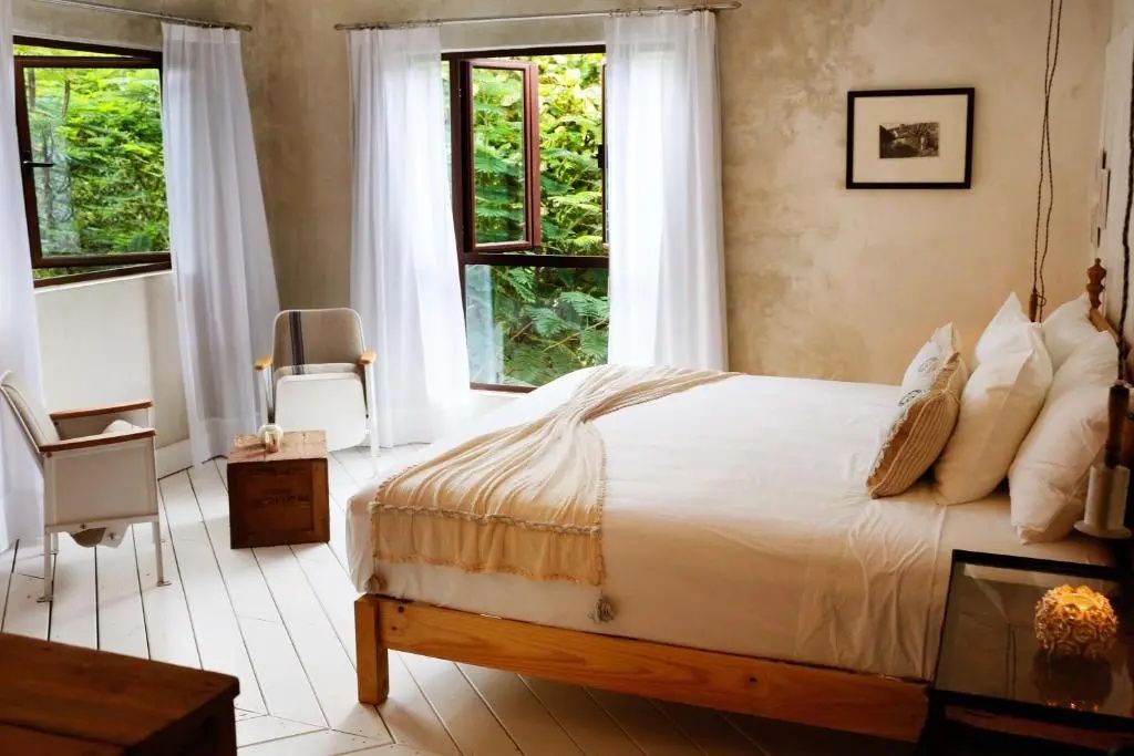 Helles Hotelzimmer mit einem Holzdoppelbett, weißer Bettwäsche und Fenster ins Grüne.