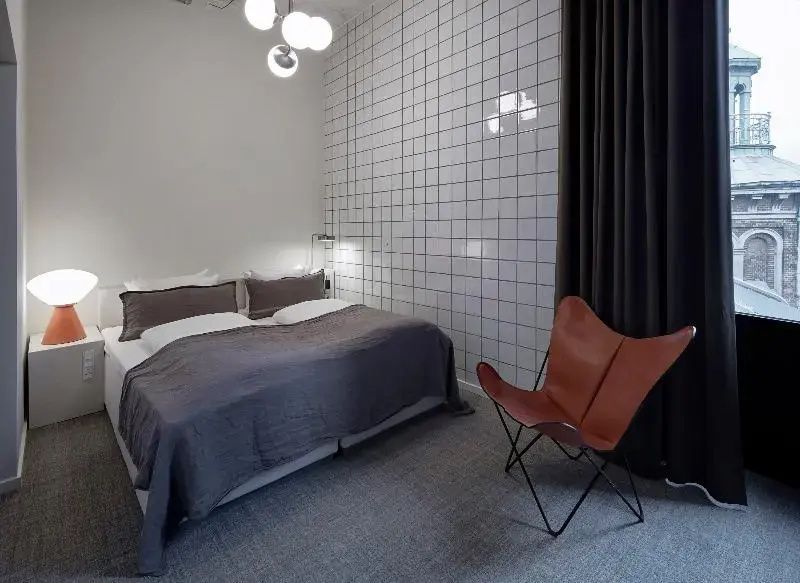 Hotelzimmer mit Doppelbett, weißer Fliesenwand, grauem Teppich und einem Ledersessel.