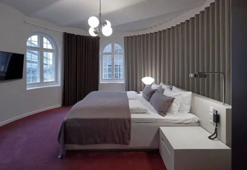 Schlafbereich mit Doppelbett, rotem Teppichboden, Rundbogenfenstern und grauem Vorhang.