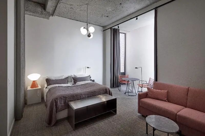 Geräumiges Hotelzimmer mit Sichtbetondecke, Doppelbett, rosa Sofa und kleiner Sitzecke.