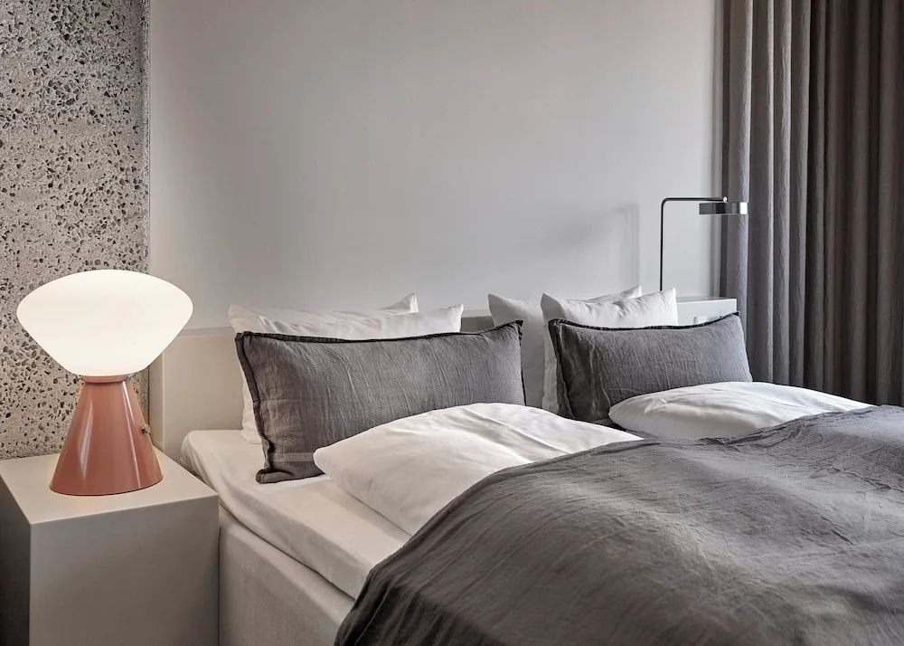 Bett mit weißen Kissen und dunkelgrauer Bettwäsche neben einer Nachttischlampe.