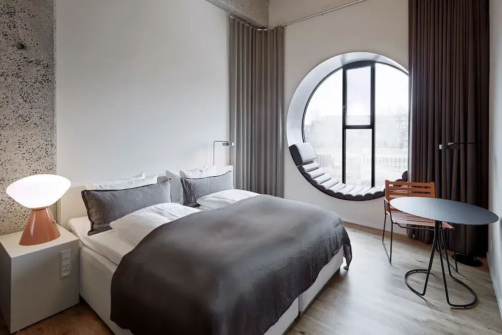 Hotelzimmer mit weißem Doppelbett und einem großen runden Fenster mit Sitznische