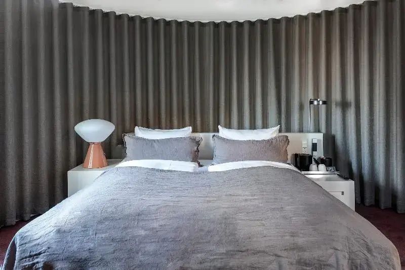 Doppelbett mit grauer Tagesdecke vor einer Strukturwand und Nachttisch mit Lampe.