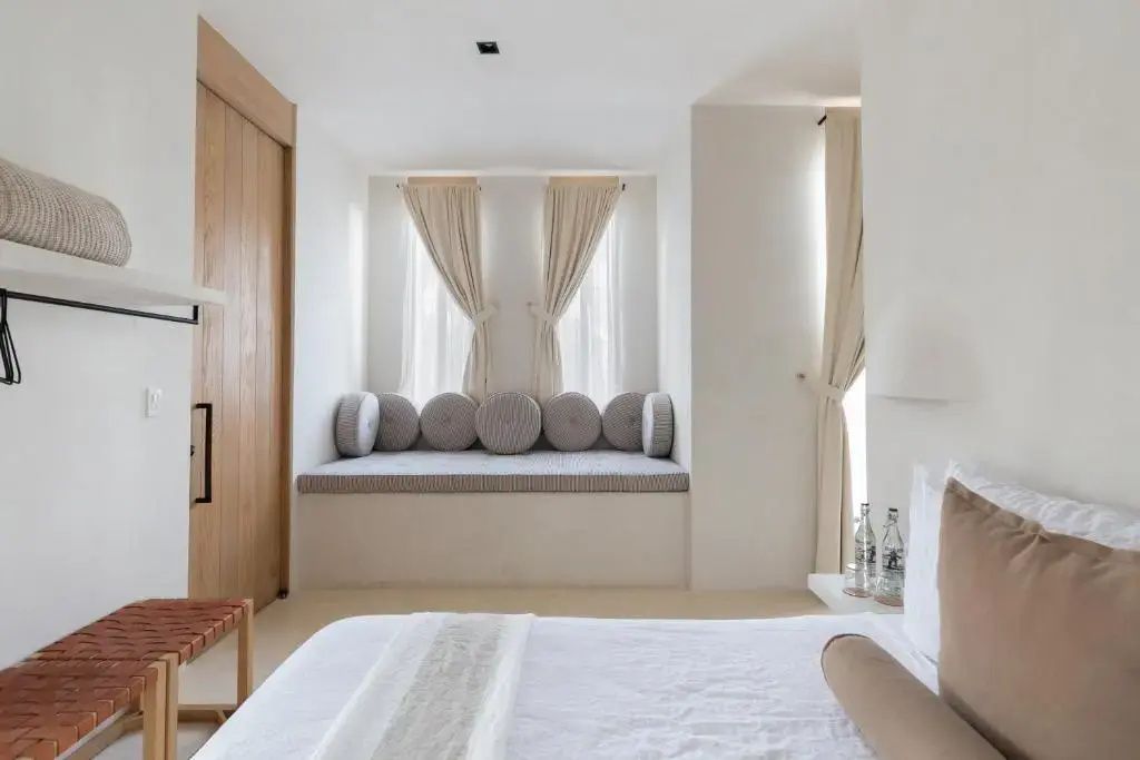 Heller Hotelraum mit weiß gemachtem Bett und gepolsterter Sitznische am Fenster.