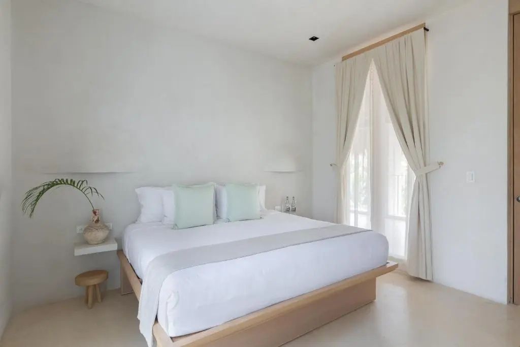 Helles Hotelzimmer mit großem Doppelbett, hellen Vorhängen und schlichter Dekoration.