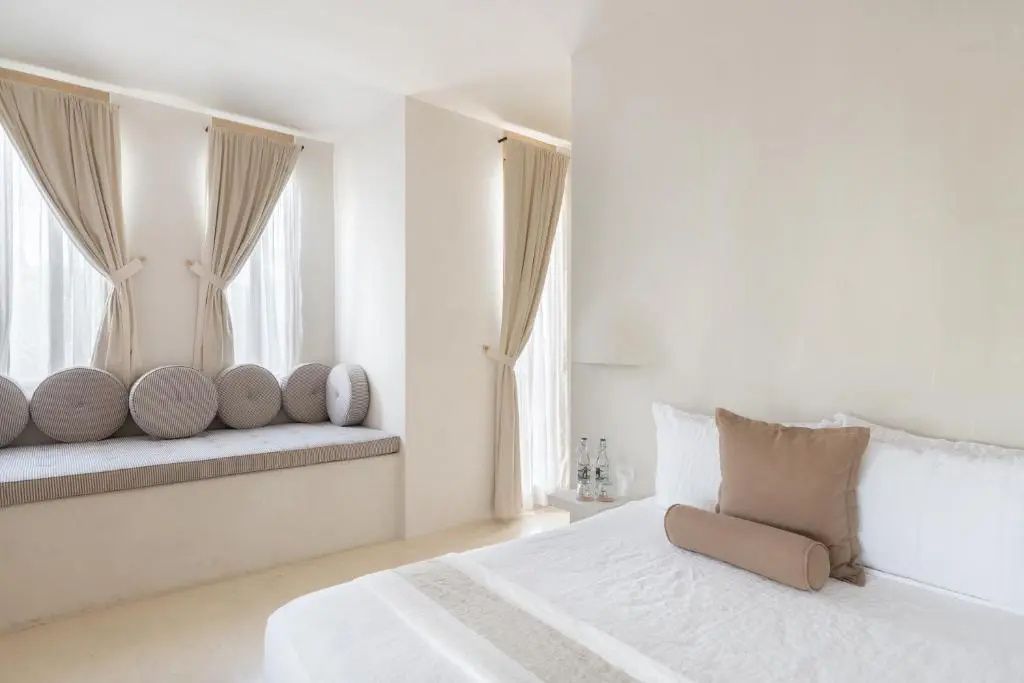 Helles Schlafzimmer mit weißem Bett, beigen Vorhängen und Sitzbank mit Rundkissen.
