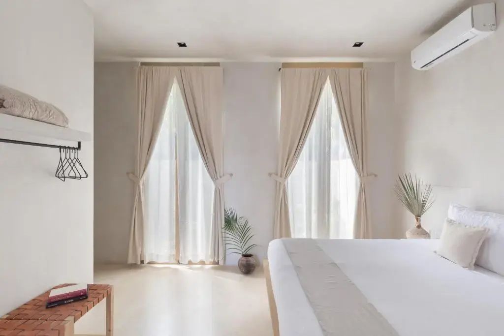 Helles Hotelzimmer mit Doppelbett, hellen Vorhängen an bodentiefen Fenstern und Zimmerpflanze.