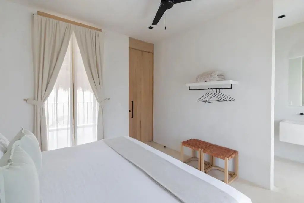 Blick über ein weißes Doppelbett auf ein Wandregal, Holzschiebetür und Deckenventilator.