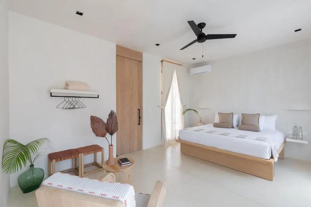 Hell gestaltetes Hotelzimmer mit Doppelbett, Deckenventilator, Sessel und einer Holztür.